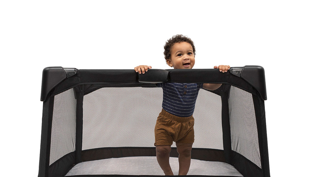 A toddler stands in a black playpen, against a white background